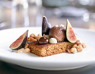 Dessert featuring a rectangular gingerbread cake topped with chocolate mousse, white chocolate chips, and garnished with fresh fig slices and candied nuts.