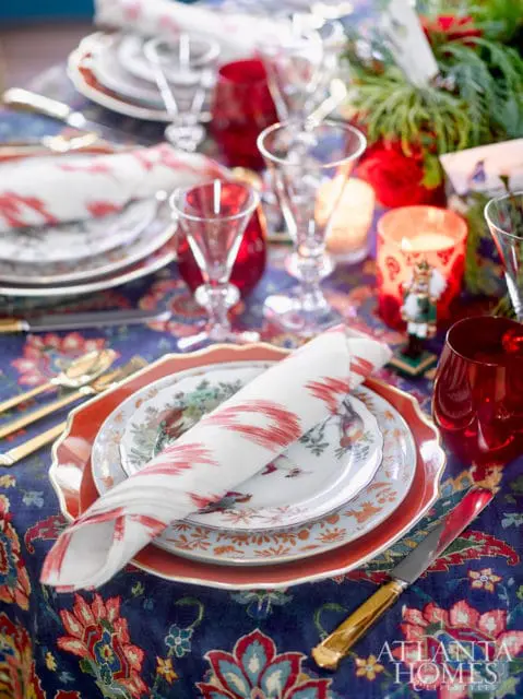 Table setting with blue paisley tablecloth, red charger plates, bird-themed china, white napkins with red stripes, gold cutlery, and red glassware.
