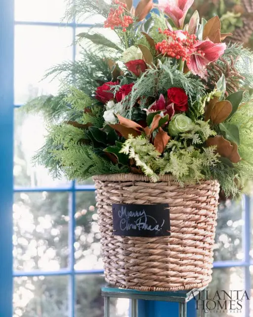 Woven basket filled with red roses, white roses, amaryllis, cedar, and magnolia leaves, with a small chalkboard tag reading "Merry Christmas!"
