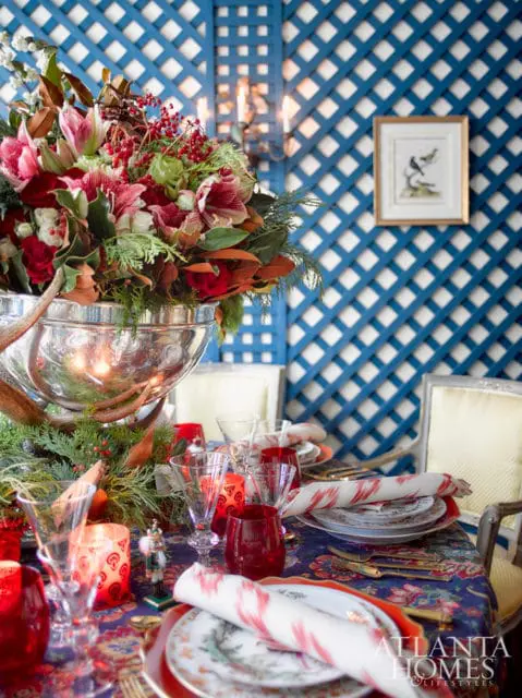 Dining table set for a holiday meal with a large floral arrangement, red glassware, nutcracker figurines, and festive patterned napkins against a blue lattice wall.