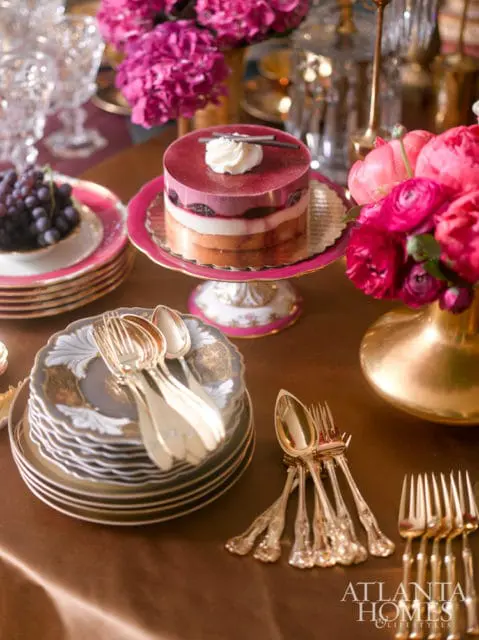 A layered berry mousse cake on a pink pedestal sits beside a stack of ornate gray and gold plates and a gold vase filled with pink flowers.
