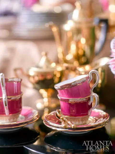 Stack of two pink teacups with gold trim and white handles, resting on saucers with gold trim. A gold teapot is visible in the blurred background.