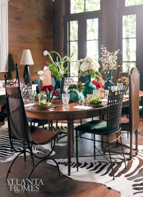 Dining room with a round wooden table set for a meal, featuring white tulips, hydrangeas, and red roses in green vases, surrounded by leather and woven chairs on a zebra print rug.