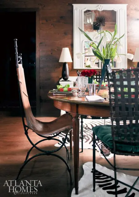 Dining room with wood-paneled walls, a round wooden table set with festive red and green wrapped gifts, white tulips in a vase, and a mix of leather butterfly chairs and dark metal chairs with green cushions.