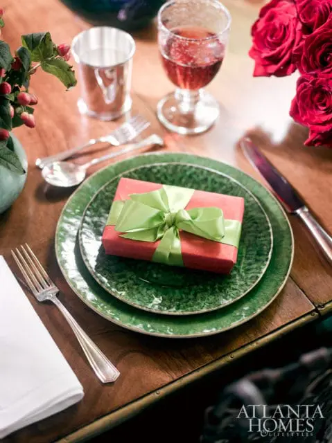 A place setting with green textured plates, a red gift box tied with a green ribbon, and silver cutlery on a wooden table.