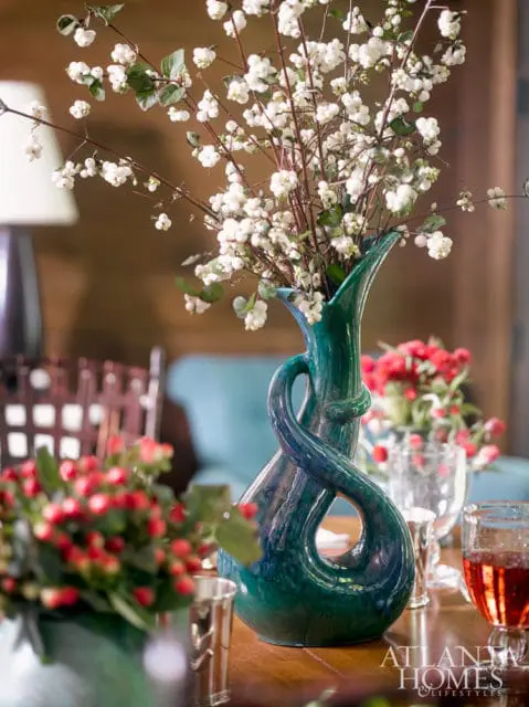A teal ceramic vase with a twisted design holds white berry branches. Red berries in a silver vase are in the foreground.
