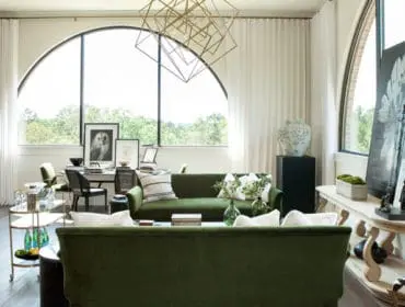 Living room with green velvet sofas, large arched window overlooking trees, and a geometric gold chandelier.