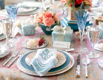 Table setting with blue crystal glasses, silver cutlery, floral patterned plates, and a monogrammed napkin.