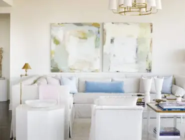 Living room with a sectional sofa, abstract art, and white furniture. Dominant colors are white and light blue, with accents of gold and brass.