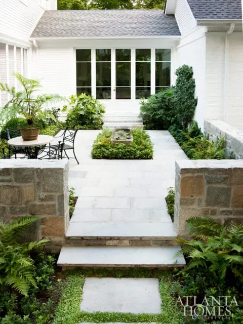 Stone pathway and steps leading to a white house with large glass doors, surrounded by lush greenery and a stone wall.