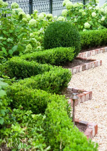 Formal garden with manicured boxwood hedges, hydrangeas, gravel path, and brick edging. A small landscape light is visible.