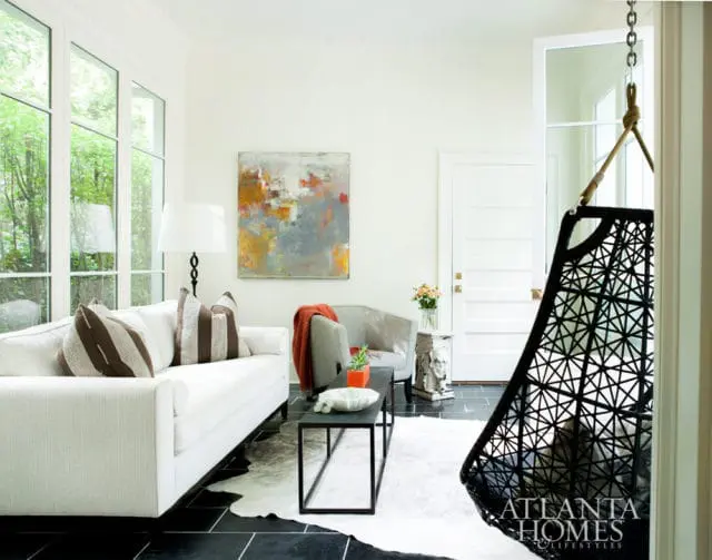 White sofa with striped pillows, abstract art, and a black hanging chair in a living room with black tile floors and large windows.