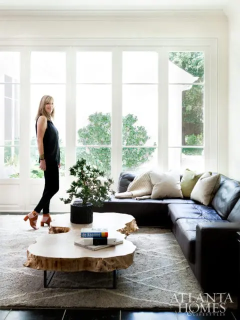 Living room with a woman standing by large windows, a dark leather sectional sofa, a live-edge wood coffee table, and a textured rug.