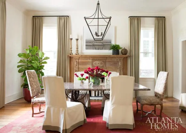 Dining room with a marble-topped table, slipcovered chairs, ornate wooden fireplace, and a large fiddle-leaf fig plant.