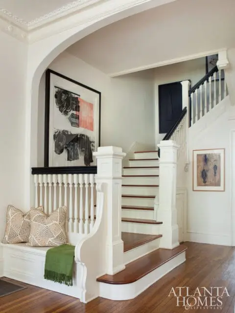 White staircase with wooden treads and risers, white balusters, and black handrail. Built-in white bench with patterned pillows and green throw sits beneath a large abstract artwork.