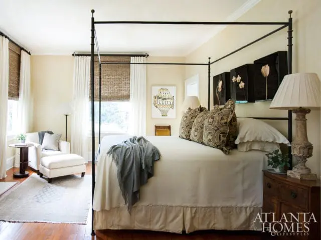 Bedroom with a black metal four-poster bed, cream bedding, patterned pillows, and a gray throw blanket. A white armchair and ottoman sit near a window with woven blinds.