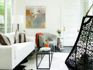 Living room with white sofa, abstract art, gray armchair with orange throw, and black geometric hanging chair.