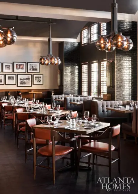 Restaurant dining room with dark wood tables, leather chairs, tufted banquettes, and globe pendant lights. Black brick walls and framed photographs adorn the walls.