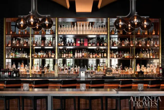 Bar with extensive liquor shelves, pendant lights, and leather bar stools. Atlanta Homes & Lifestyles logo in the lower right corner.