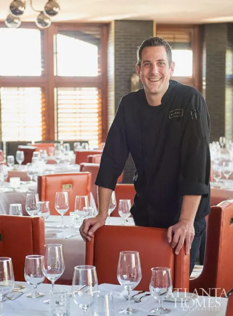 Chef in a black uniform smiling and leaning on an orange chair in a restaurant dining room with tables set for dinner.