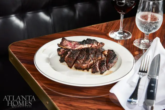 Sliced steak with dark crust on a white plate with "AMERICAN CUT" text, next to a glass of red wine and a glass of sparkling water on a wooden table.