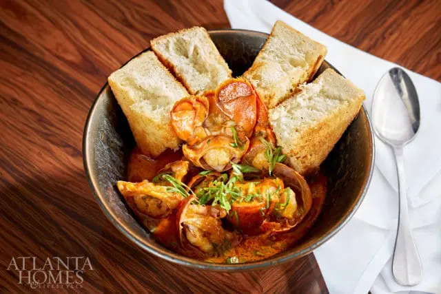 Bowl of lobster stew with toasted bread slices and garnished with green herbs, served on a wooden table with a white napkin and spoon.