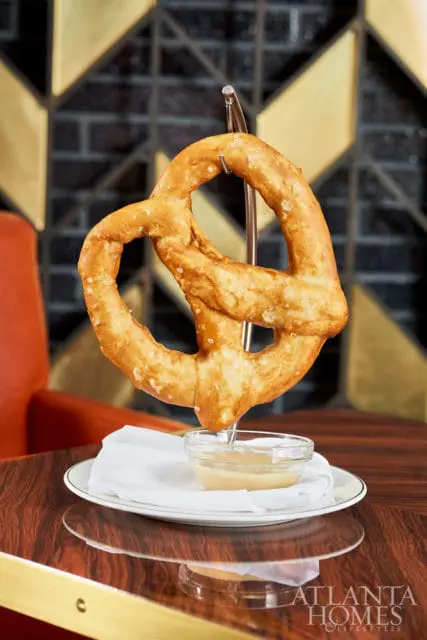 Large pretzel suspended over a small glass bowl of dipping sauce, with a white napkin and plate on a polished wood table.