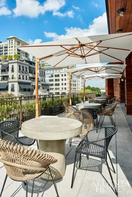Outdoor patio seating area with concrete tables and rattan chairs, shaded by white umbrellas, overlooking city buildings.
