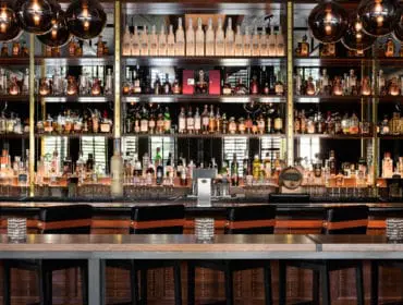 Bar with extensive liquor shelves, mirrored back wall, and pendant lighting. A long wooden bar top with black stools with orange accents is in the foreground.