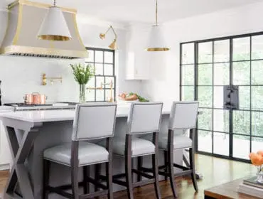 White kitchen with a large brass range hood, gray island with white marble countertop, and light gray upholstered bar stools. Large black-framed windows overlook greenery.