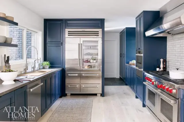 Navy blue kitchen cabinets with stainless steel appliances including a large refrigerator and a double oven range with red knobs. White subway tile backsplash above the range.
