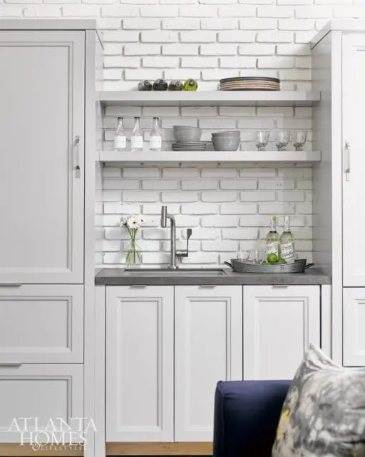 White brick backsplash in a kitchen with light gray cabinetry, a dark gray countertop, and open shelving displaying dishes and bottles.