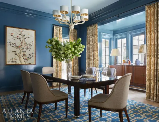 Blue dining room with dark wood table, upholstered chairs, patterned curtains, and a painting of birds on a tree