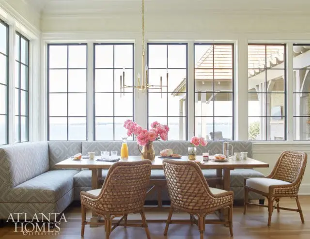 Breakfast nook with a U-shaped banquette upholstered in patterned fabric, a wooden table set for two, and rattan chairs. Large windows overlook a body of water.