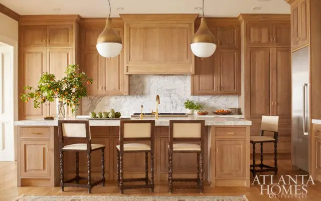 Warm wood kitchen with marble countertops, brass fixtures, and pendant lighting. Features a large island with seating and a stainless steel refrigerator.