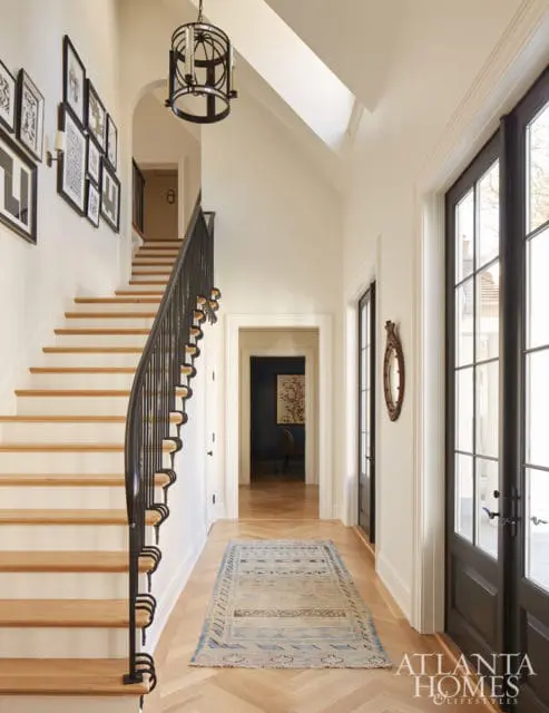 Hallway with a curved staircase featuring light wood treads and a black metal railing, a patterned rug on a herringbone wood floor, and a large black-framed door.