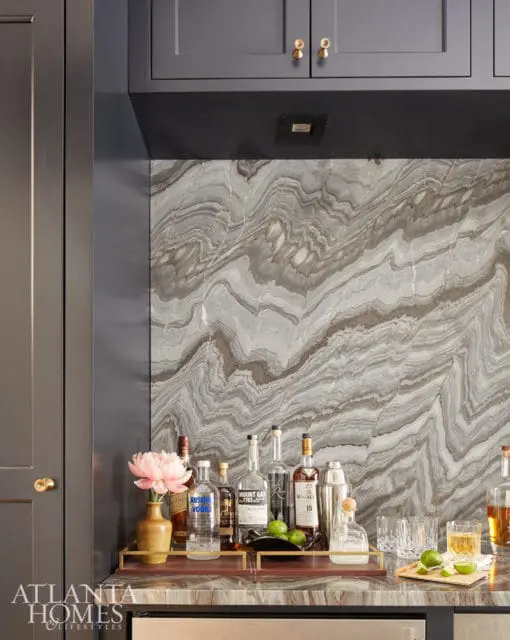 Bar area with dark gray cabinetry, a dramatic swirled marble backsplash, and a collection of liquor bottles on a marble countertop.