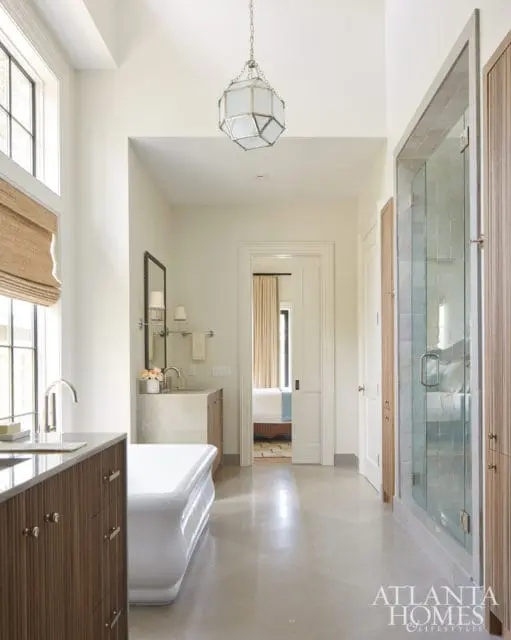 Modern bathroom with a freestanding white tub, glass-enclosed shower, and wood-paneled cabinetry. A doorway leads to a bedroom.