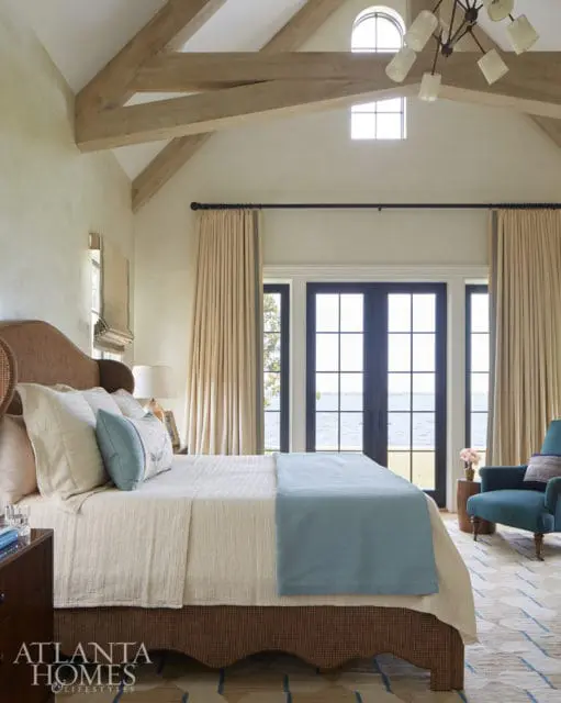 Bedroom with vaulted ceiling, exposed wooden beams, and large windows overlooking a body of water. Features a bed with light-colored linens and a blue throw, a teal armchair, and a patterned rug.