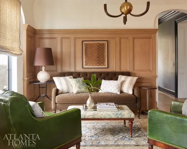 Living room with wood-paneled walls, a brown tufted sofa, green leather armchairs, and a patterned ottoman.
