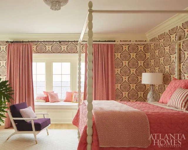 Bedroom with a four-poster bed, coral bedding, patterned wallpaper, and coral curtains. A window seat with pillows overlooks the water.