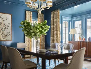 Blue dining room with dark wood table, upholstered chairs, patterned curtains, and a painting of birds on a tree branch.