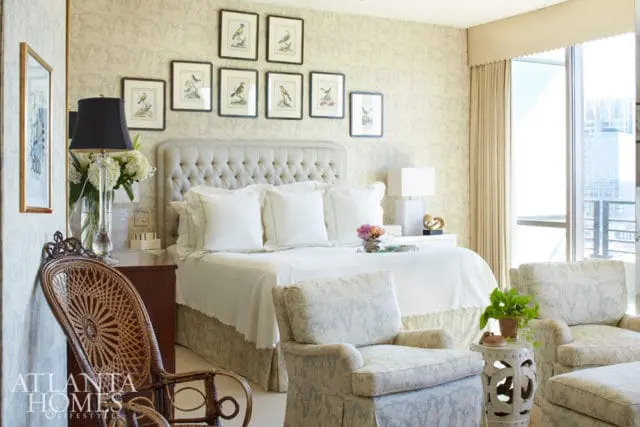 Bedroom with a tufted headboard, white bedding, and patterned wallpaper. A wicker chair and two upholstered armchairs are in the foreground.