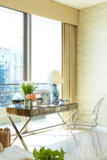 Mirrored desk with chrome legs, featuring a lamp, flowers, and decorative items, paired with a clear acrylic chair in a room with patterned wallpaper and large windows.