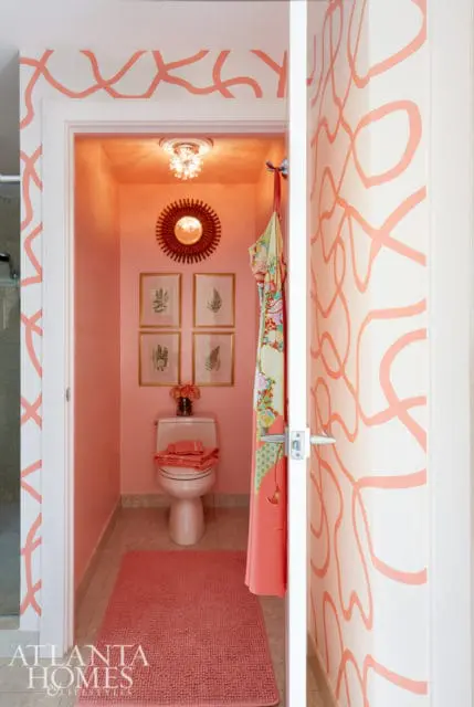 Pink bathroom with coral abstract wallpaper, a sunburst mirror, framed botanical prints, and a pink rug in front of the toilet.