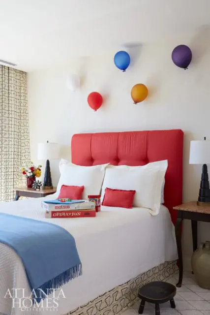 Bedroom with a red tufted headboard, white bedding, and colorful balloons on the wall. Monopoly and Operation board games are stacked on the bed.