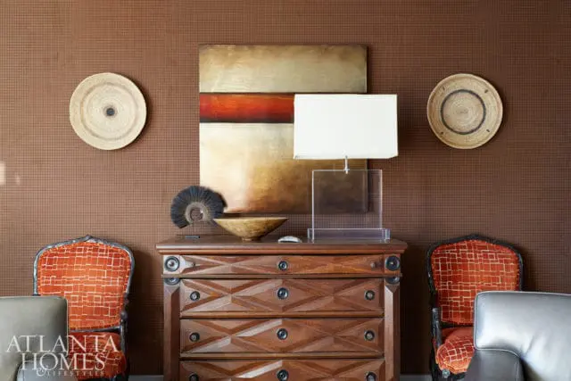 Brown textured wallpaper adorns a wall above a wooden dresser with geometric carvings. Two chairs with orange patterned upholstery flank the dresser, which holds a lamp, artwork, and decorative objects.
