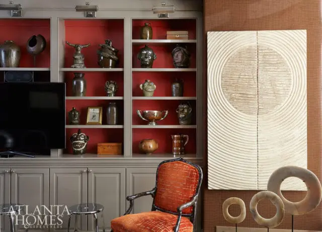 Built-in gray cabinetry with red shelves displaying various ceramic jugs, pottery, and decorative objects. An orange upholstered armchair sits in front of the shelves.