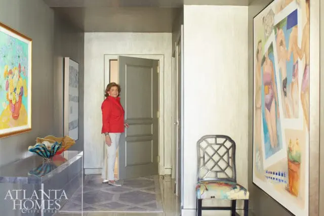 Woman in red jacket and white pants stands in a hallway with gray walls, a gray door, and geometric patterned floor. Artwork adorns the walls.