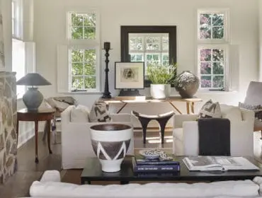 Living room with stone fireplace, neutral-toned sofas, wooden console table with decorative objects, and multiple windows overlooking greenery.